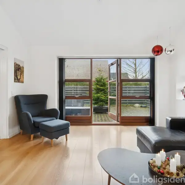 Stue med sort lædersofa og lænestol står i et lyst rum med trægulv, hvorfra en dør leder ud til en terrasse med træer udenfor. Bord med hvide lys i forgrunden.