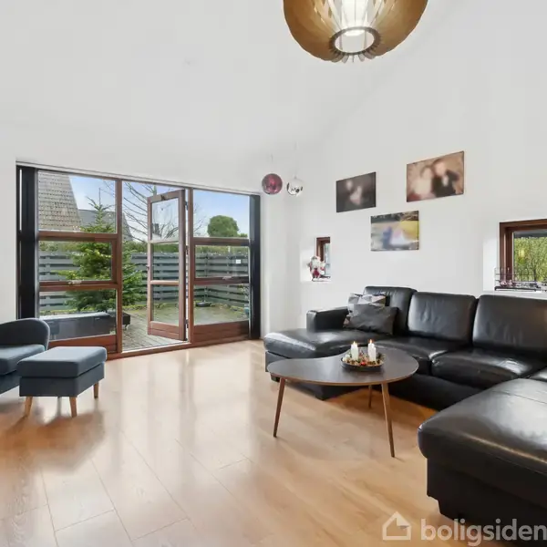 Et moderne rum med en sort lædersofa i et hjørne, sofabord foran, og lænestol med skammel ved store vinduer, der åbner mod en terrasse. Væggene har enkelte billeder.