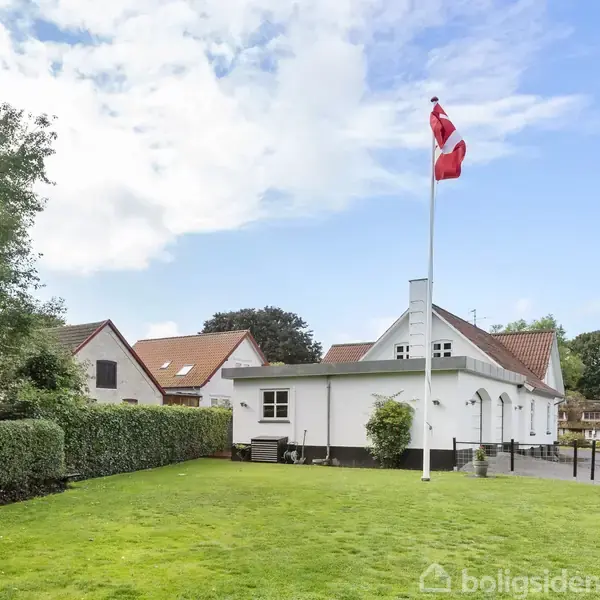 Et hvidt hus med rødt tegltag står ved en velplejet grøn græsplæne. En dansk flagstang pryder haven, omgivet af hække og træer under en delvist skyet himmel.