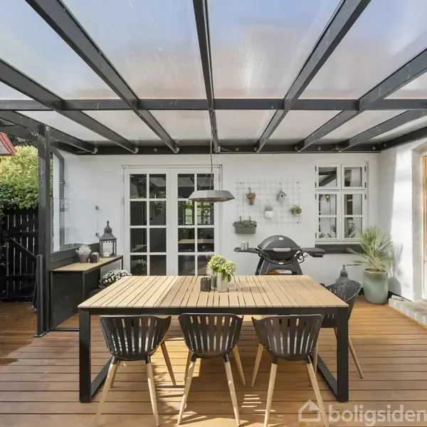 En træterrasse med et stort spisebord og fire stole står under et gennemsigtigt tag. En kuglegrill er til højre, omringet af planter mod en hvid husmur med døre og vinduer.