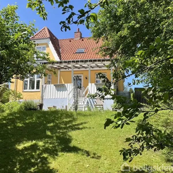 Gult hus med rødt tag står omgivet af træer og en grøn have. En trappe fører op til hoveddøren, og der er en veranda foran huset. Solrigt vejr.