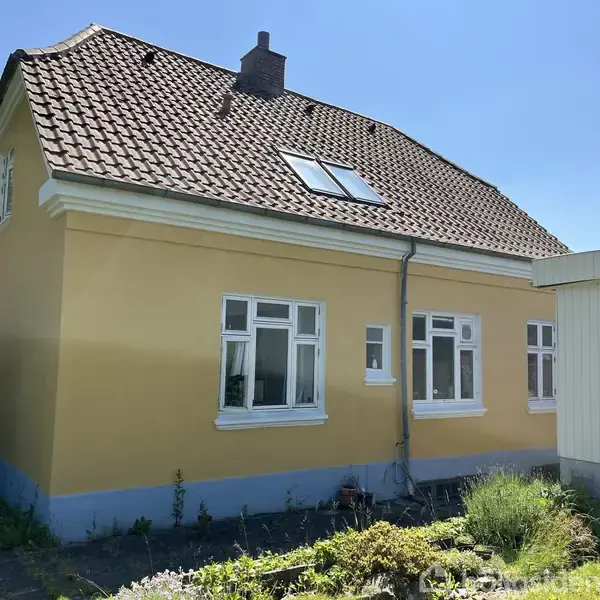 Gult hus med rødt tegltag og loftsvindue står i en have. Vinduerne er hvide, og solen skinner klart.