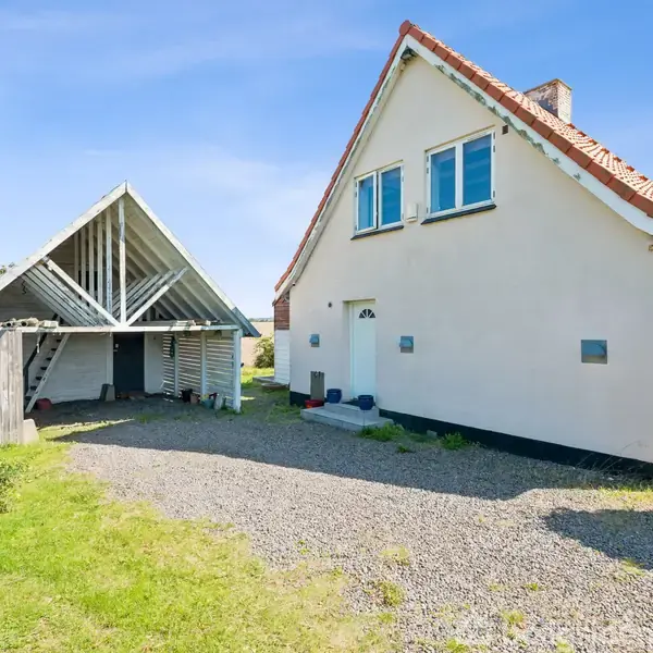 Hus med hvid facade og rødt tag står ved siden af en åben carport. Omgivet af grusbelagt indkørsel og grønne marker under en klar blå himmel.