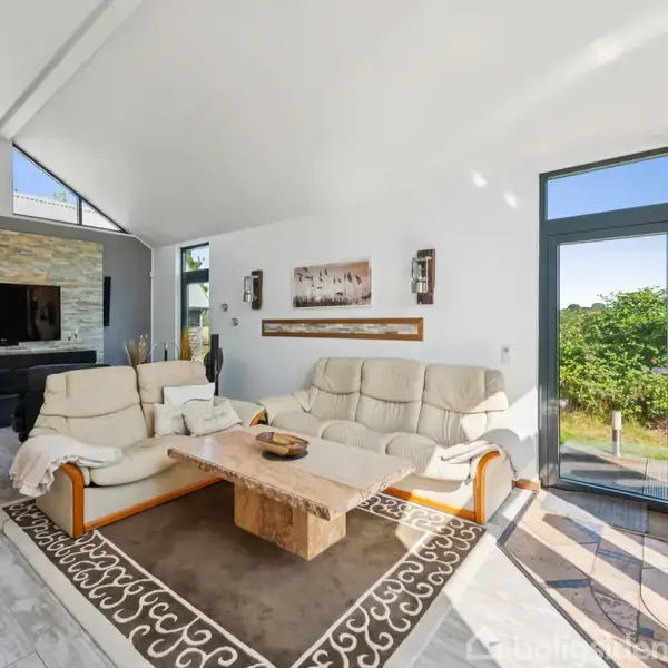 En moderne stue med lyse lædersofaer, der står omkring et træbord. I baggrunden hænger et fladskærms-tv på en stenvæg. Store vinduer giver udsigt til grøn natur. Pejsen til højre er tændt.