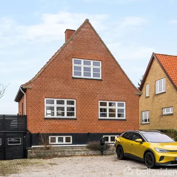 Rød murstensvilla med sadeltag og hvide vinduer står stille ved siden af en gul bil. Foran huset er en grusbelagt parkeringsplads, og nabohuset har også rødt tegltag.