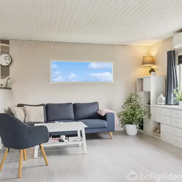 En stue med en blå sofa og lænestol ved et sofabord, placeret foran et vindue med udsigt. Til højre er en reol med planter og en lampe.