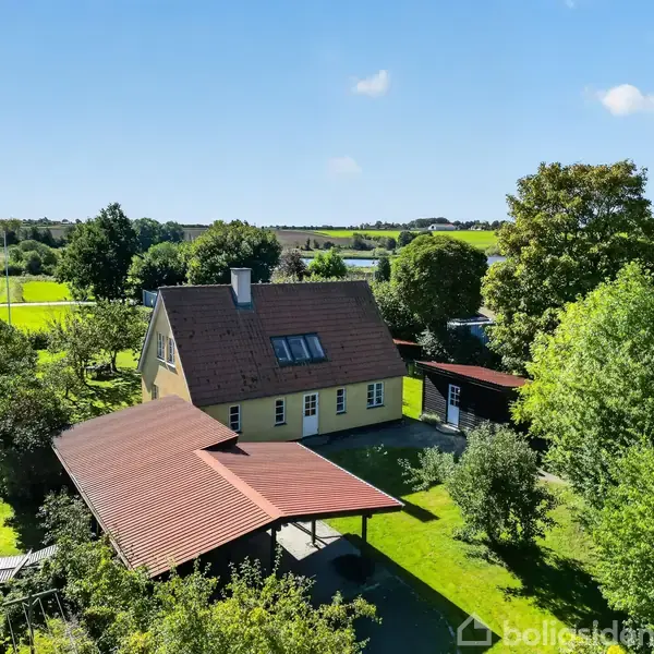 Et gult hus med rødt tag står i en frodig have med træer og buske. En carport med rødt tag ses foran, omgivet af grønne marker og landskab.