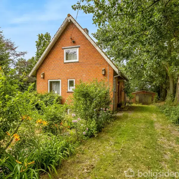Rødt murstenshus står stille blandt frodig vegetation. Grussti fører væk fra huset, omgivet af træer og blomster. Ingen synlig tekst.