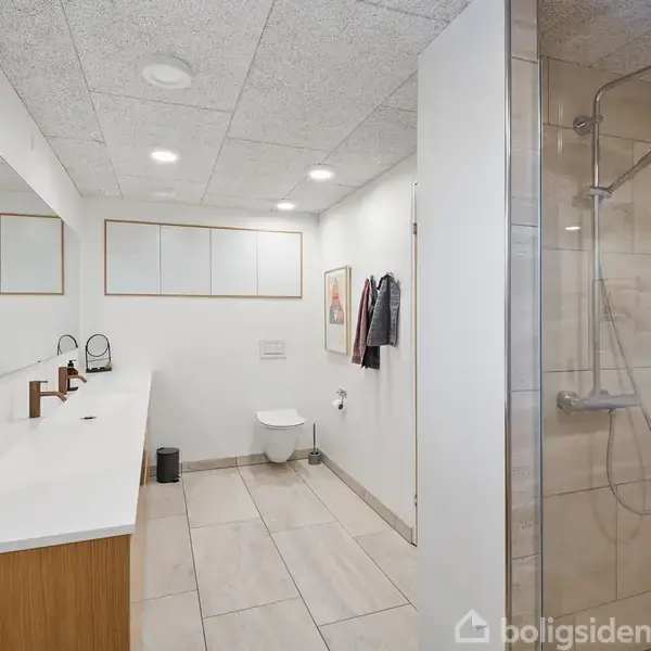 Badeværelse med dobbelthåndvask i hvid bordplade, brusekabine til højre. Vægophængt toilet foran stor vægmonteret spejl. Loft med indbyggede loftslamper. Rene og minimalistiske linjer.