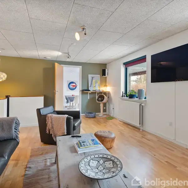 Sort lædersofa med gråt tæppe står i hjørnet af en stue. Et lille træbord foran sofaen har en dekorativ skål. Til højre hænger et fladskærms-tv på væggen. I baggrunden ses et åbent rum med en dør og flere kunstgenstande på væggen. Lyse trægulve og moderne indretning.