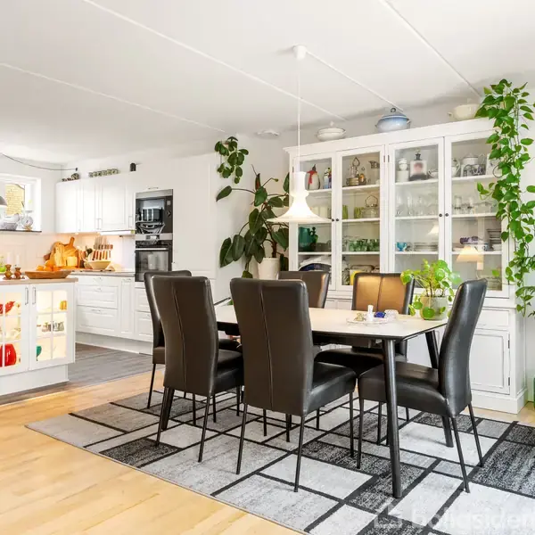 En spisebord omgivet af sorte stole står på et tæppe i en stue-køkken-alrum. Hvidt vitrineskab fyldt med glasvarer og planter i baggrunden. Rummets stil er lys og moderne.