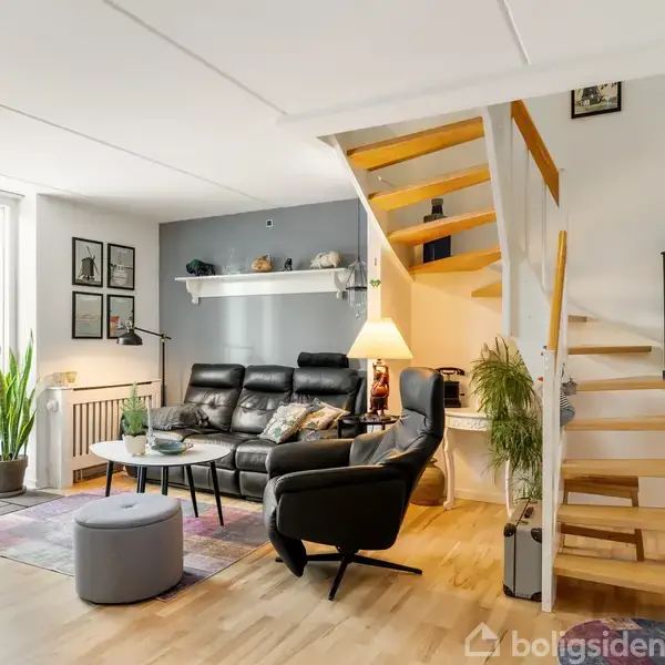 Sort lædersofa står foran hvid væg under trappe i stue. Trætrappen fører opad. Store vinduer bader rummet i lys, grønne planter giver liv. Hyggelige lamper og billeder skaber varme.