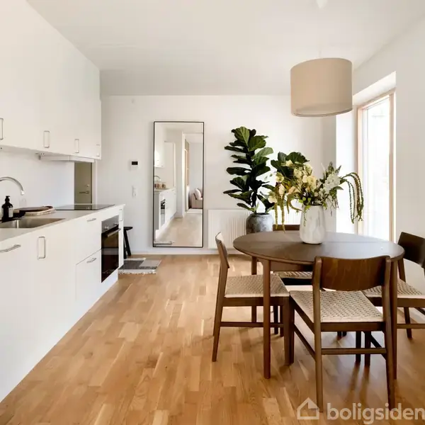 Et moderne køkken-alrum med en hvid køkkensektion til venstre. Spisebord med fire stole står til højre, dekoreret med en vase blomster. Store vinduer giver dagslys.