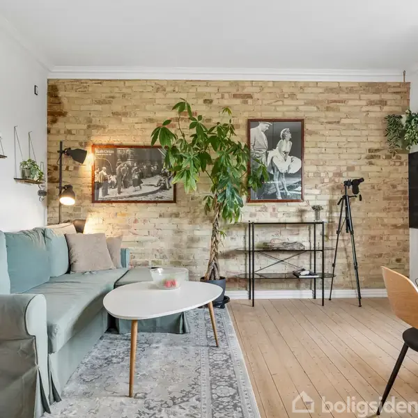 Stue med grøn sofa ved siden af et rundt sofabord. En murstensvæg med plante, billeder og en hylde. Et kamera står på stativ ved siden af et stort TV.