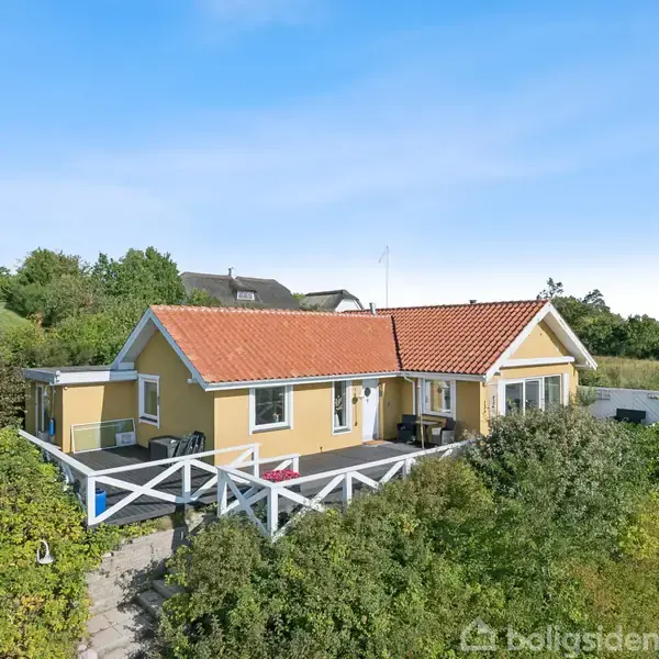 Gult hus med rødt tag står omgivet af frodig have. Det har en stor terrasse med hvidt gelænder, placeret i et naturskønt, grønt område under blå himmel.