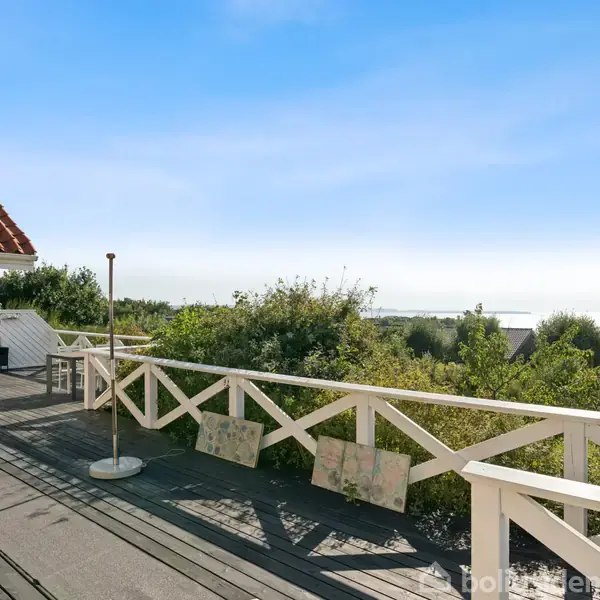En terrasse med hvidt rækværk, hvor to stole står udenfor et gult hus. I baggrunden ses grønt løv og udsigt over havet.