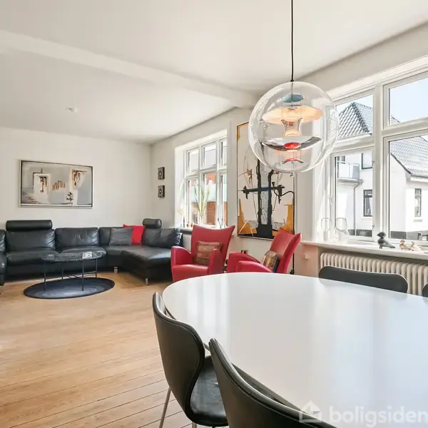 En moderne stue med en rund sort sofa og to røde lænestole placeret foran store vinduer. Rummet har trægulv og et hvidt spisebord med sorte stole.