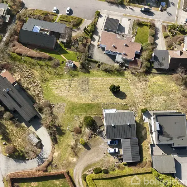 Boligområde set fra oven med huse placeret langs veje og grønne græsarealer. Der er flere indkørsler, og biler ses parkeret ved nogle huse.