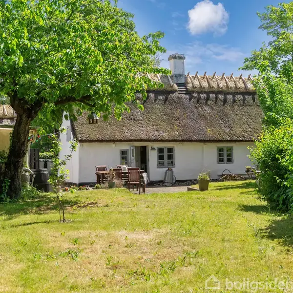 Et bindingsværkshus med stråtag står i en frodig have. Havemøbler er placeret på en terrasse foran huset, omgivet af grønne buske og et stort træ.