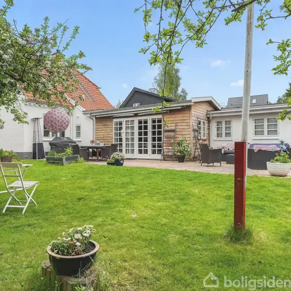 En hvid murstenshus med en træbeklædt terrasse hvor et sæt havemøbler er placeret. Omkranset af en frodig have med grønne træer og græsplane.
