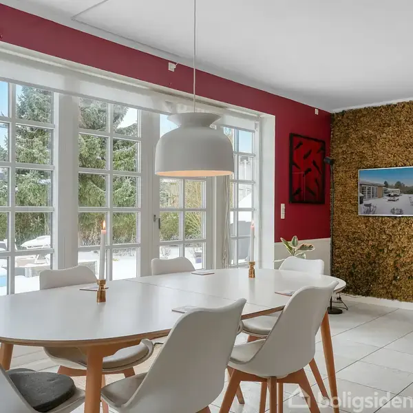 Spisebord omgivet af hvide stole står i et rum med rød og hvid væg. En loftlampe hænger over bordet. Store vinduer giver udsigt til en solrig have. En vægskærm viser et billede.