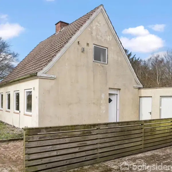 Et lille, ældre hus med skråt tag står i et åbent område omgivet af træer. Foran huset er der et hegn og en have.