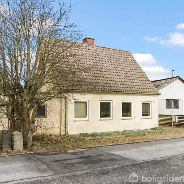 Et hvidt, en-etagers hus med brun tagstenstag står på en gade, omgivet af en større træ ved siden af og en anden bygning i baggrunden.