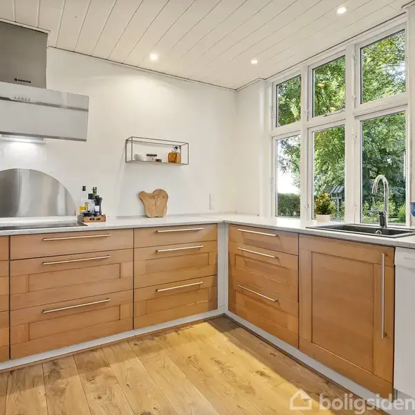 Køkken med træskabsfacader og en hvid bordplade omkranset af store vinduer. Køkkenvasken er placeret til højre, og emhætte hænger over kogepladen. Udsigt til grøn have.