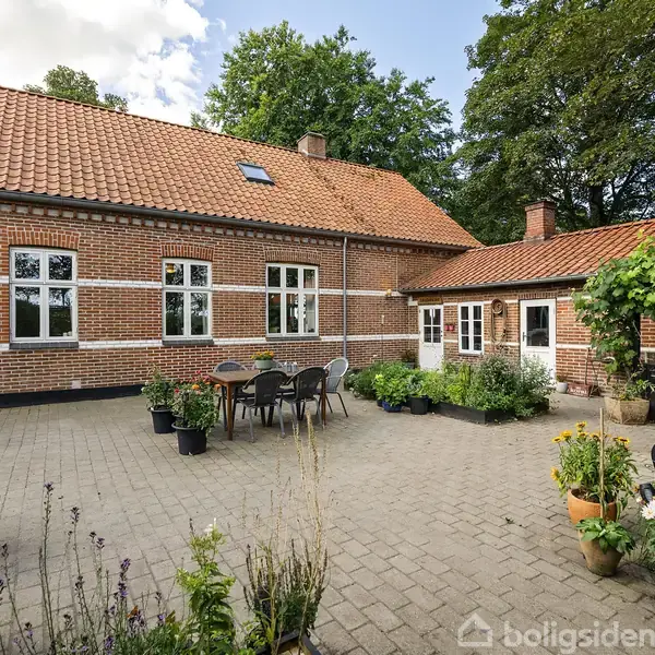 Et rødt murstenshus med rødt tegltag omgiver en brostensbelagt gårdhave med havemøbler og blomsterkrukker. Omgivet af grønne træer og klar himmel.
