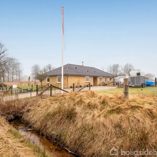Et gult murstenshus står stille i en landlig omgivelser omgivet af marker og træer. En flagstang ses foran huset ved siden af en grusvej og en lille bæk.