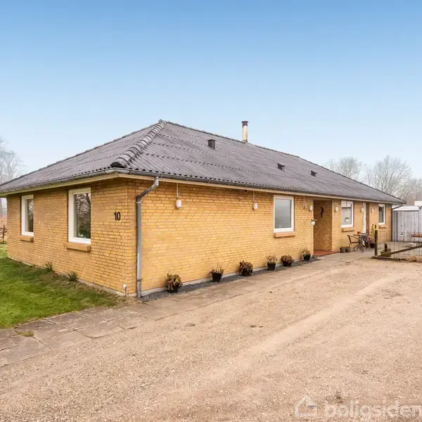 Et gult murstenshus med sort tegltag står stille i et åbent, grønt landskab. Der står "10" på siden af huset, og der er en grusbelagt indkørsel foran.