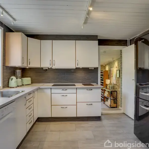 Et hvidt køkken med skabsmoduler og apparater, inklusive ovn og køleskab, i et lyst rum. Rummet har et stort vindue med udsigt til grønne træer udenfor.