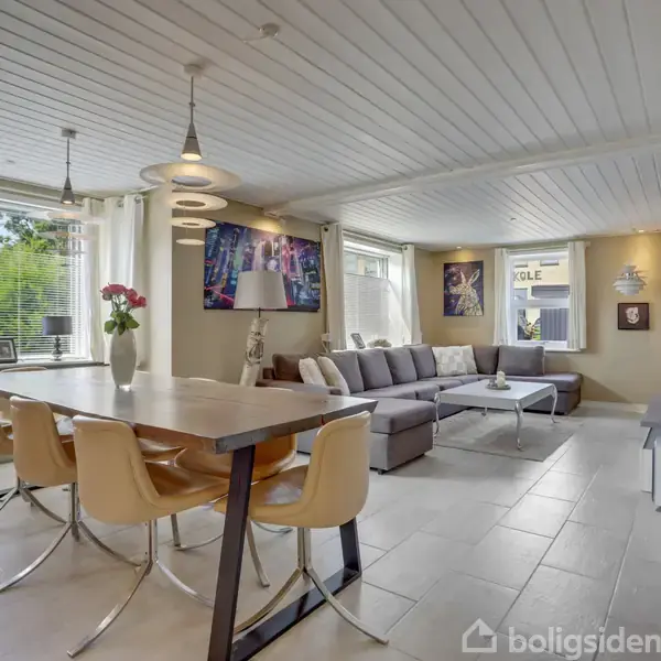 Et lyst rum indeholder et stort spisebord med stole til venstre og en hjørnesofa foran et TV til højre. På væggene hænger kunstværker, og store vinduer giver udsigt til haven.