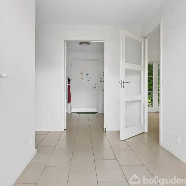 En lys gang har åbne døre og lyse flisegulve. En fjern dør viser gardiner og vægpynt. Væggene er hvide, og der ses naturligt lys fra højre.