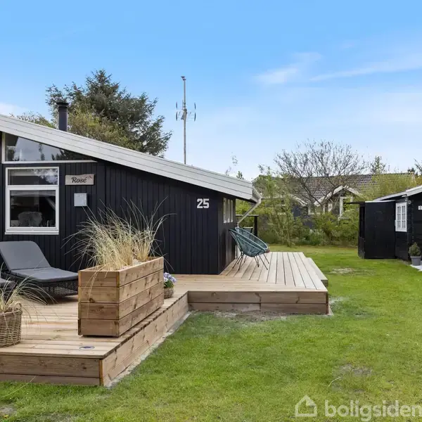 Sort træhus med nummeret 25, ligger ved en terrasse med liggestole og plantekasser. Det er omgivet af en grøn have med et mindre, sort skur i baggrunden.