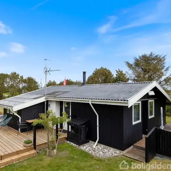 Et sort træhus står stille i en grøn have med en træterrasse. Der er havemøbler foran huset, og en gynge ses i baggrunden. Himlen er blå.