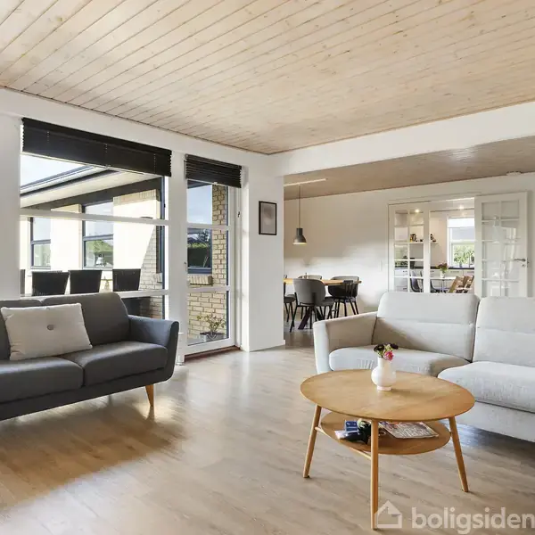 Stue med moderne sofaer foran et sofabord. Store vinduer lukker lys ind. Baggrunden viser et åbent køkken-område med spisebord og stole. En rød skur kan ses udenfor.