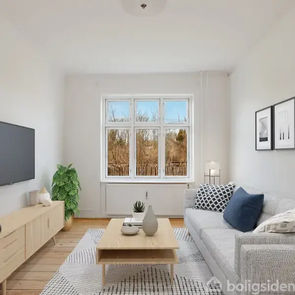 Stue med grå sofa og puder, sofabord og fladskærms-TV på væggen. Et vindue med udsigt til træer og minimalistisk kunst på væggen. Trægulv med tæppe og grøn plante.