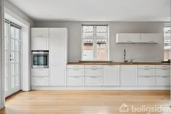 Køkken med hvide skabe og indbygget ovn placeret mod en væg. Vinduer frigiver naturligt lys ind i rummet med trægulv og enkel indretning.