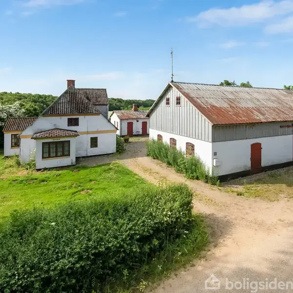 Et hvidt bondehus med rødt tag står på en grusvej ved siden af en lade. Omgivet af frodigt grønt landskab og blå himmel.