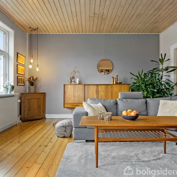 Stue med gråt hjørnesofaarrangement, træbord med skål af appelsiner, og vægdekorationer. Trægulv og loft, store planter i hjørnet, to vinduer med lysindfald, og moderne belysning.