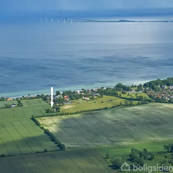 Huse med marker strækker sig ud til en kystlinje. En pil peger på et specifikt hus. Havet og en vindmøllepark ses i baggrunden.