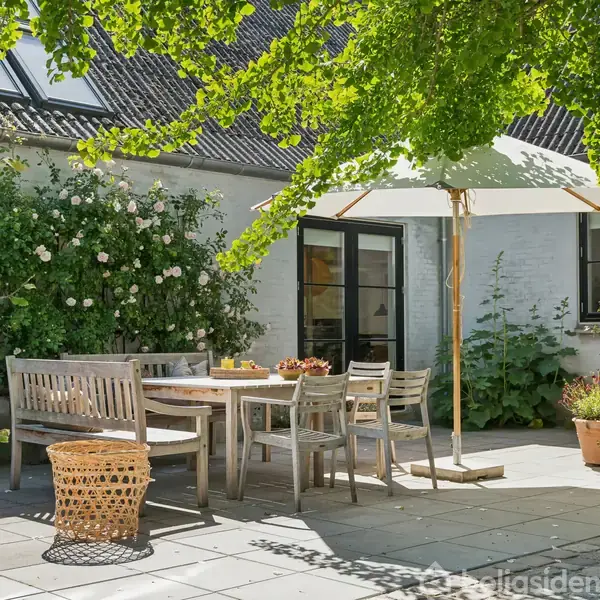 En træterrassemøbler er placeret omkring et bord under en stor parasol. Omgivet af blomstrende buske og grønne planter på en flisebelagt terrasse foran et hvidt murstenshus.