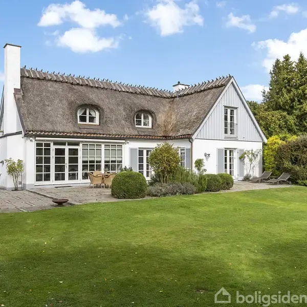 Hus med stråtag står fredeligt i en velplejet have omgivet af buske og træer under en klar blå himmel. Terrassen foran huset inviterer til udendørs ophold.