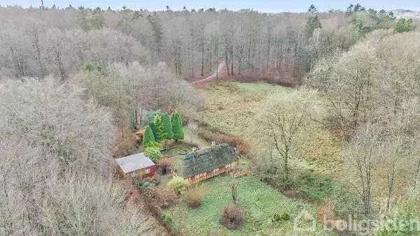 Et bindingsværkshus med stråtag omgivet af træer og en græsplæne i en skov. En lille bygning står ved siden af huset. Skovområder omgiver stedet.