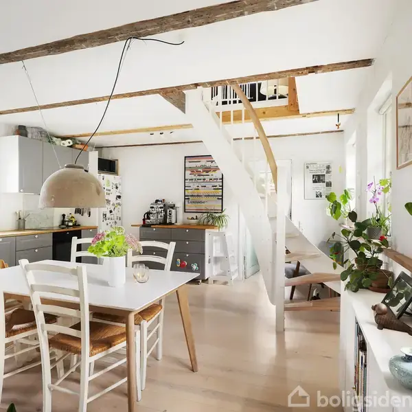 Hvidt spisebord med fire stole står i et lyst køkken, hvor planter dekorerer rummet. Trappen i baggrunden leder til en hems. Lyse træbjælker pryder loftet.