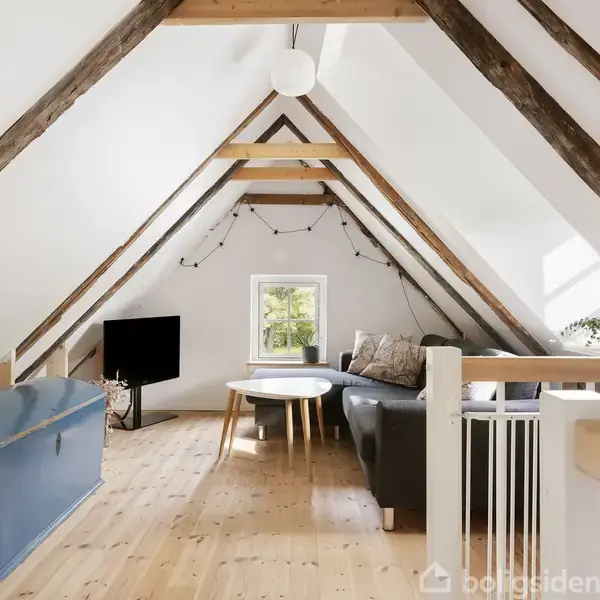 Loftstue med skrå træbjælker i loftet; en grå sofagruppe står ved et lille vindue; et rundt bord er placeret i midten; der er en fladskærms-tv.