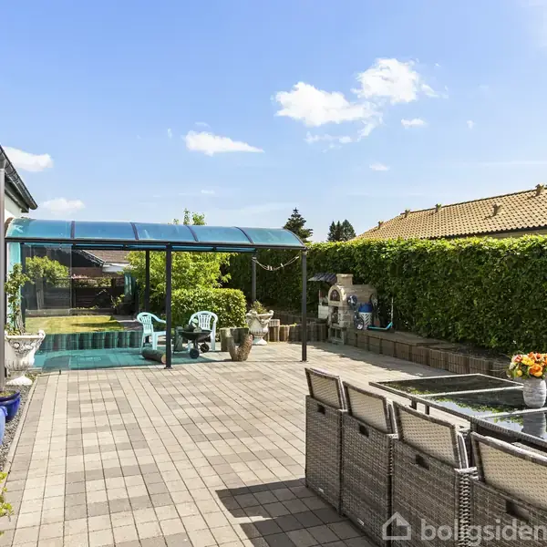 En møbleret terrasse med spisebord og stole står ved siden af en swimmingpool med overdækning, omgivet af en høj, grøn hæk. Det er en lys og solrig dag.