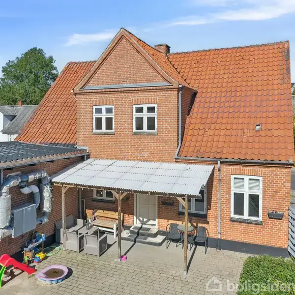 Murstenshus med rød tag, overdækket terrasse foran. Legetøj og møbler på terrassen. Nabohuse i baggrunden. Klar himmel og træer omgiver boligen.