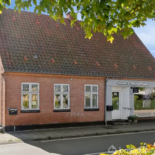 Et rødt murstenshus med sort tag står ved en vej. Skiltet over indgangen lyder "Vinne & Harry Hansen". Grønne træer omkranser scenen, og himlen er blå.